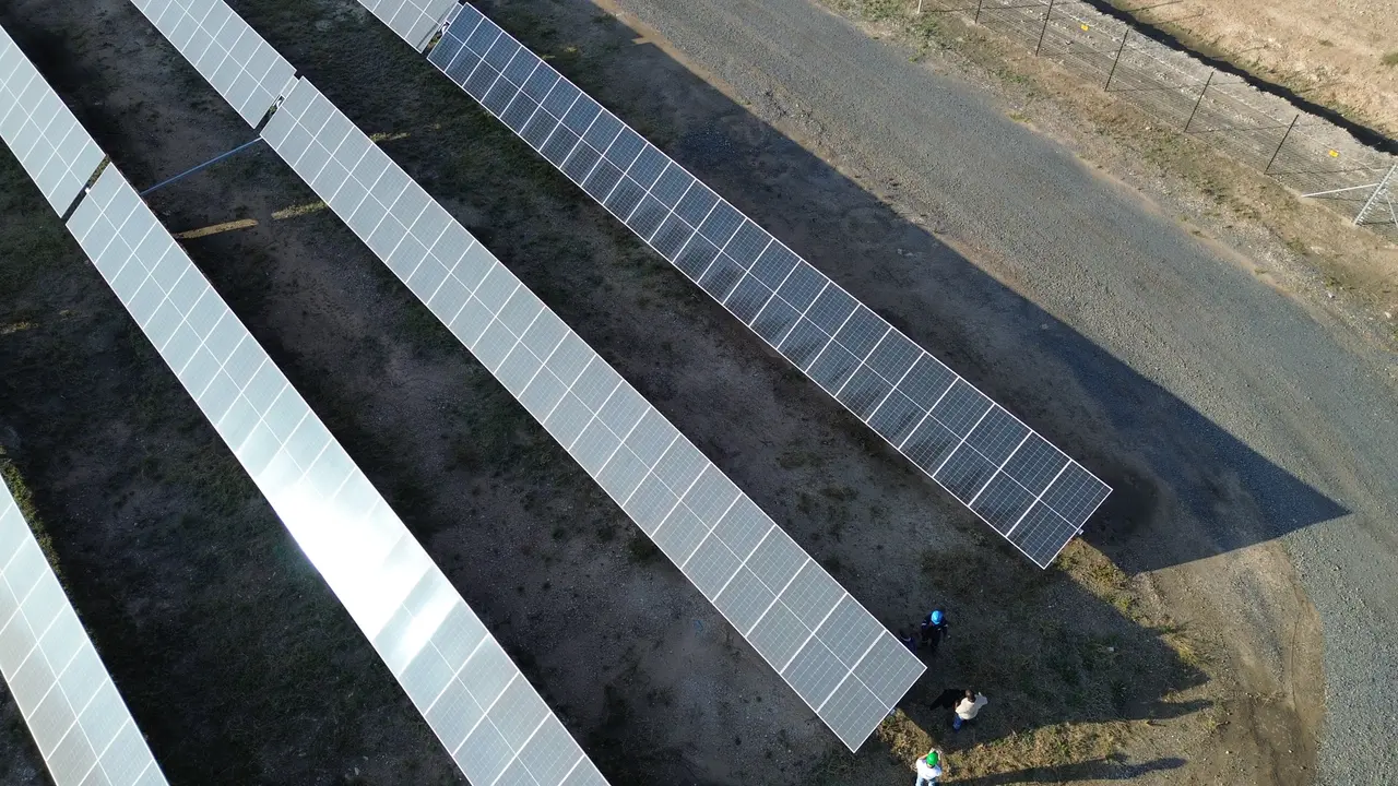 Angled drone view of grey, dust-covered module rows and one cleaned dark row, with three crew members at the end of a row