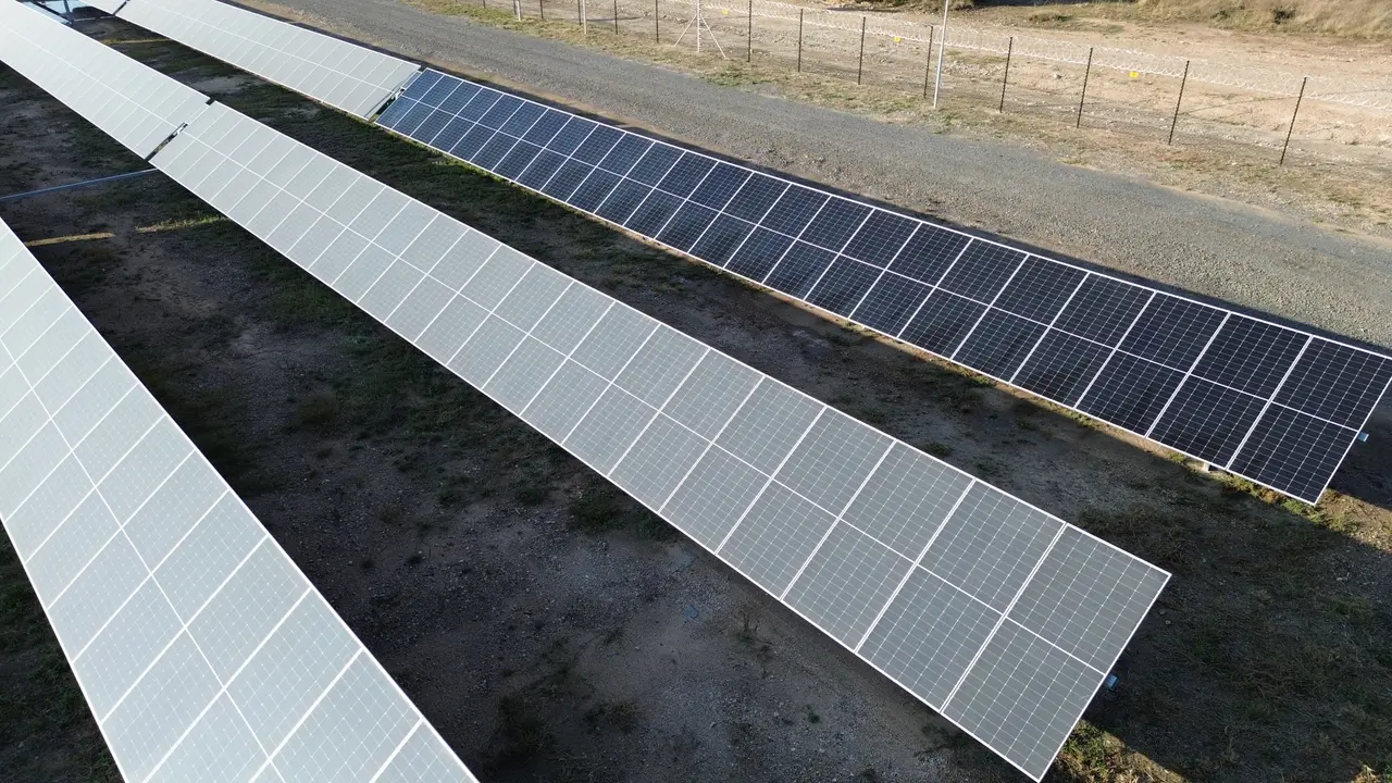 Drone view at the North West pilot site: one freshly cleaned, dark module row beside grey, dust-covered rows, next to the perimeter road and fence