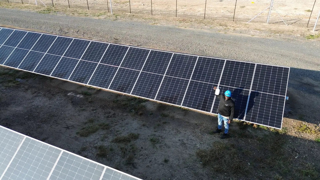 Drone view of a technician in a hard hat holding up a small handheld device beside a cleaned, dark module row, with a dusty row in the foreground