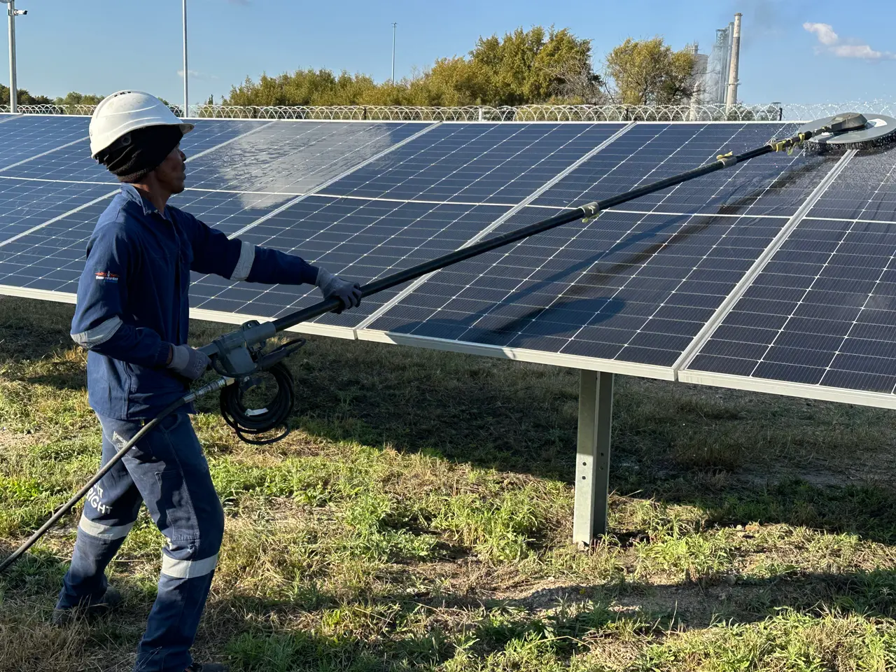 A crew member in a hard hat cleans a module row with a long-handled, water-fed rotating brush